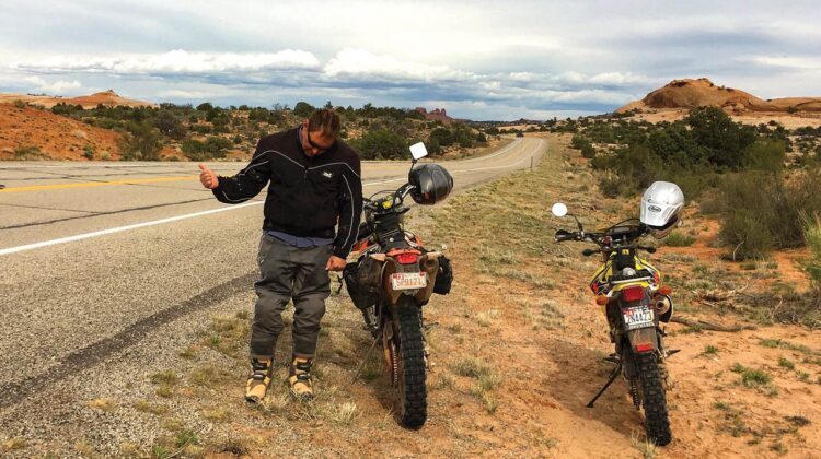 A man with his thumb extended stands beside a desert highway