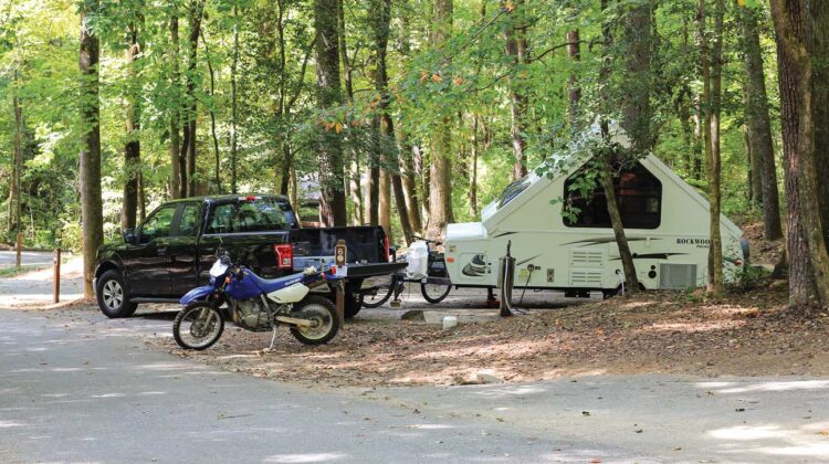 A pickup with an RV is parked in a campsite alongside a motorcycle