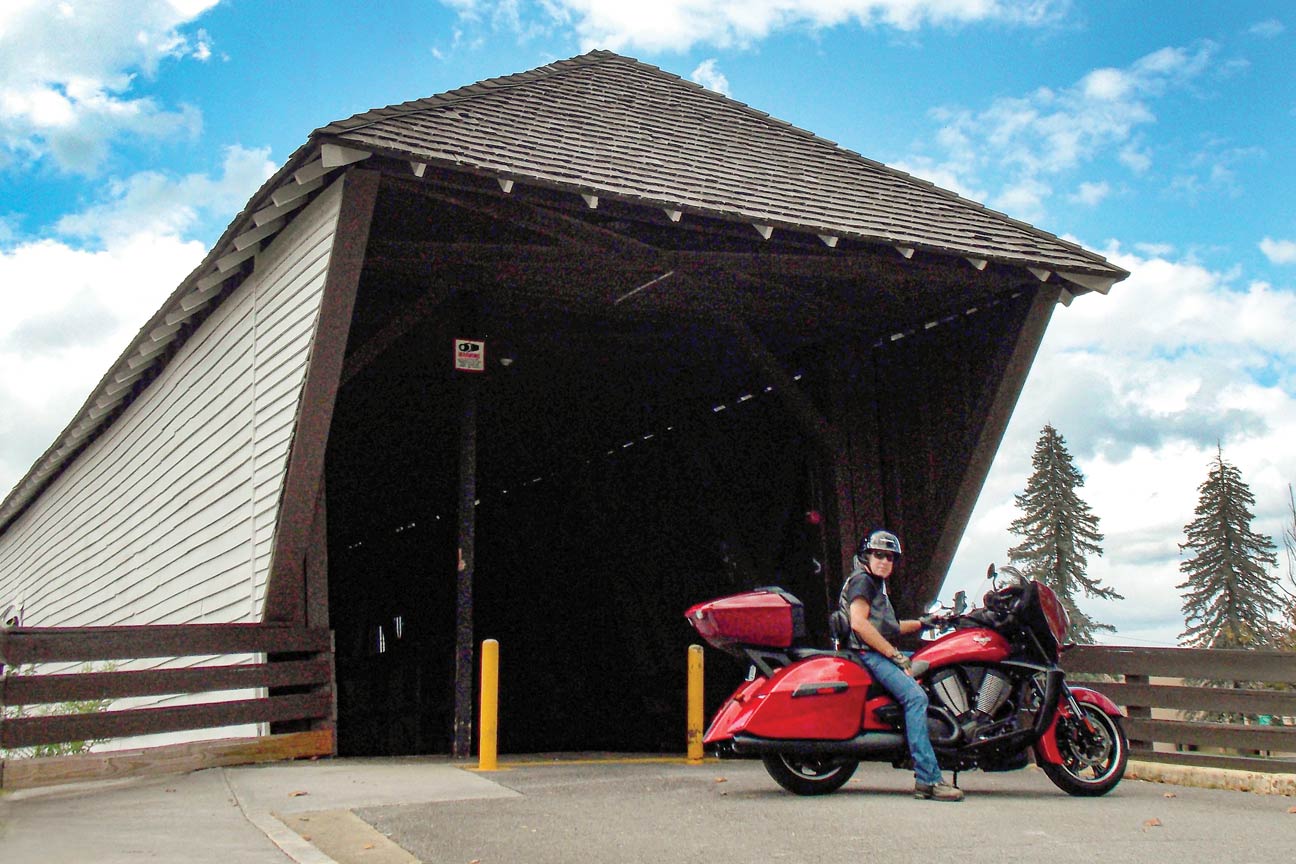 A man sits astride a red motorcycle at the entrance of a covered bridge