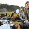 A woman stands beside her BMW motorcycle. A farm is in the background.