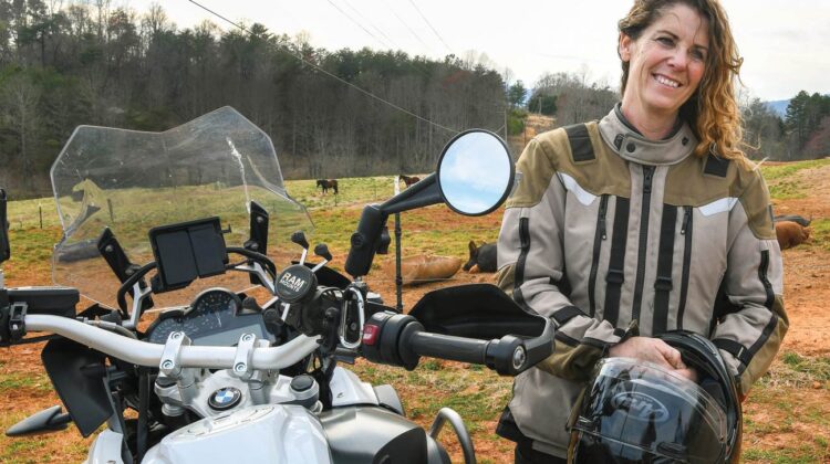 A woman stands beside her BMW motorcycle. A farm is in the background.