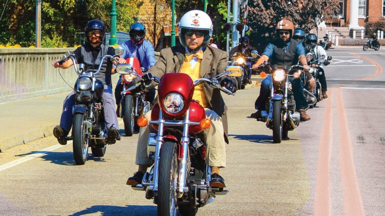 Well-dressed men ride motorcycles across a bridge