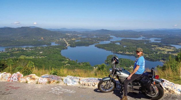 A woman sits astride her motorcycle at an overlook with a scenic view in the background.