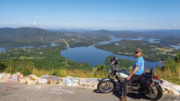 A woman sits astride her motorcycle at an overlook with a scenic view in the background.