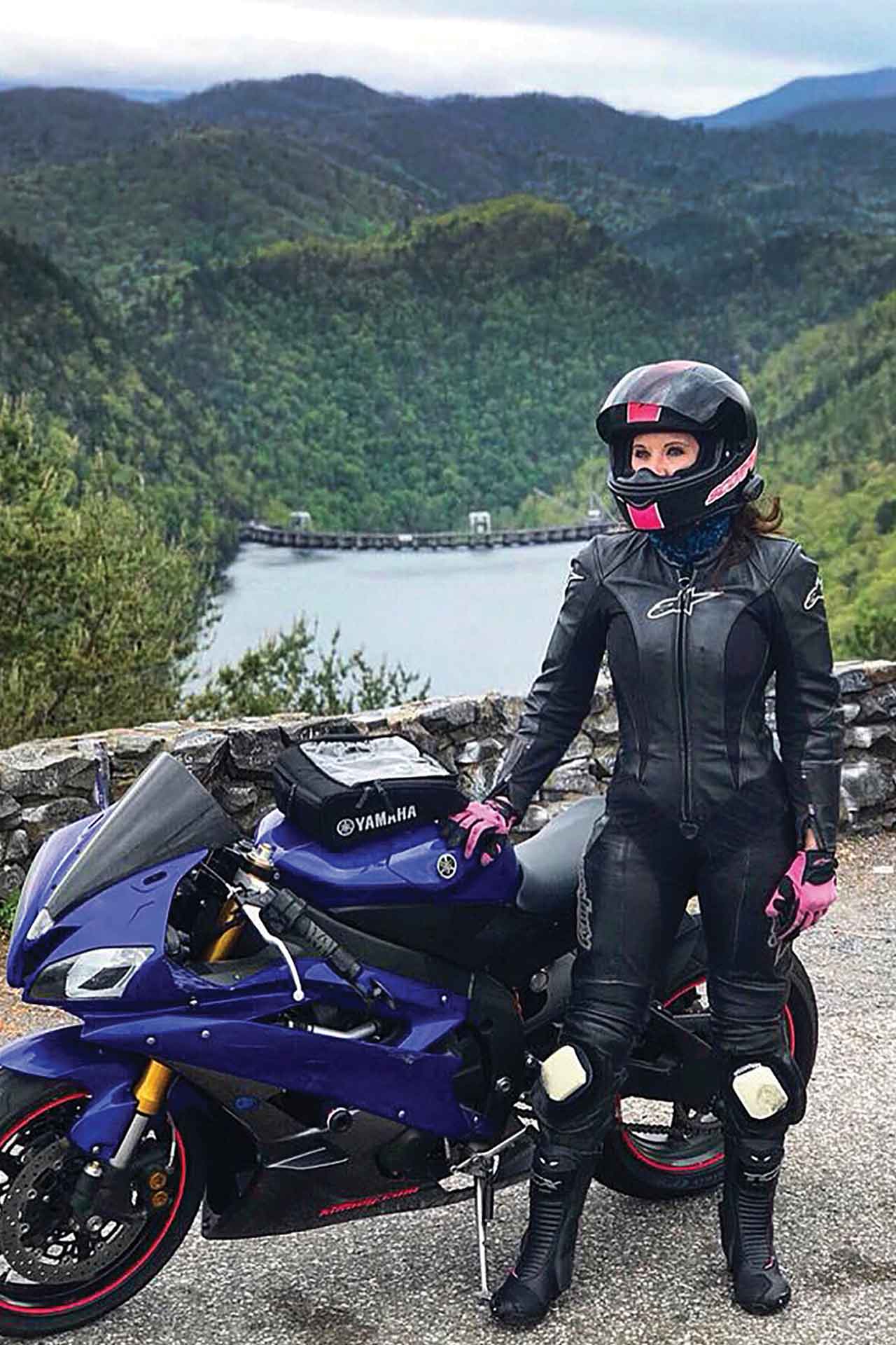 A woman at an overlook stands beside her motorcycle, a lake and dam is in the background.