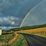 A rainbow arches over a scenic country road surrounded by a barn and golden fields