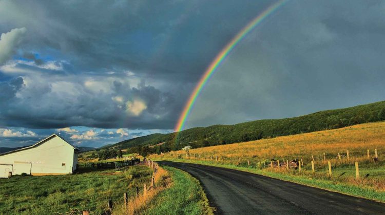 A rainbow arches over a scenic country road surrounded by a barn and golden fields