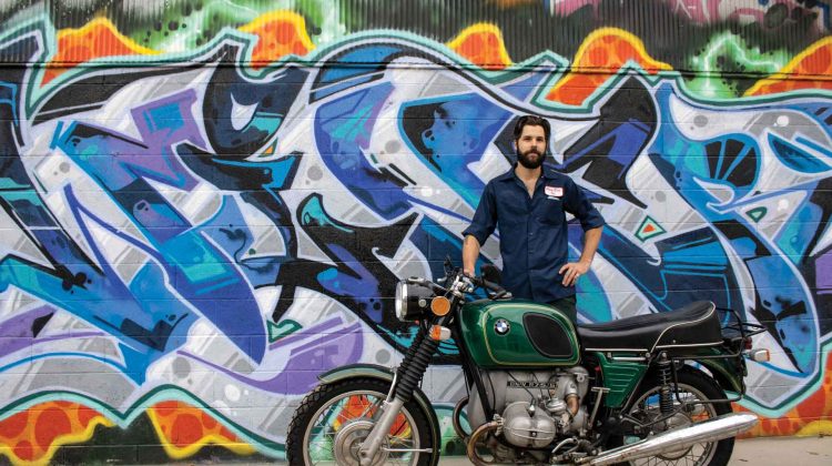 A man stands with a vintage BMW motorcycle in front of a colorful graffiti mural