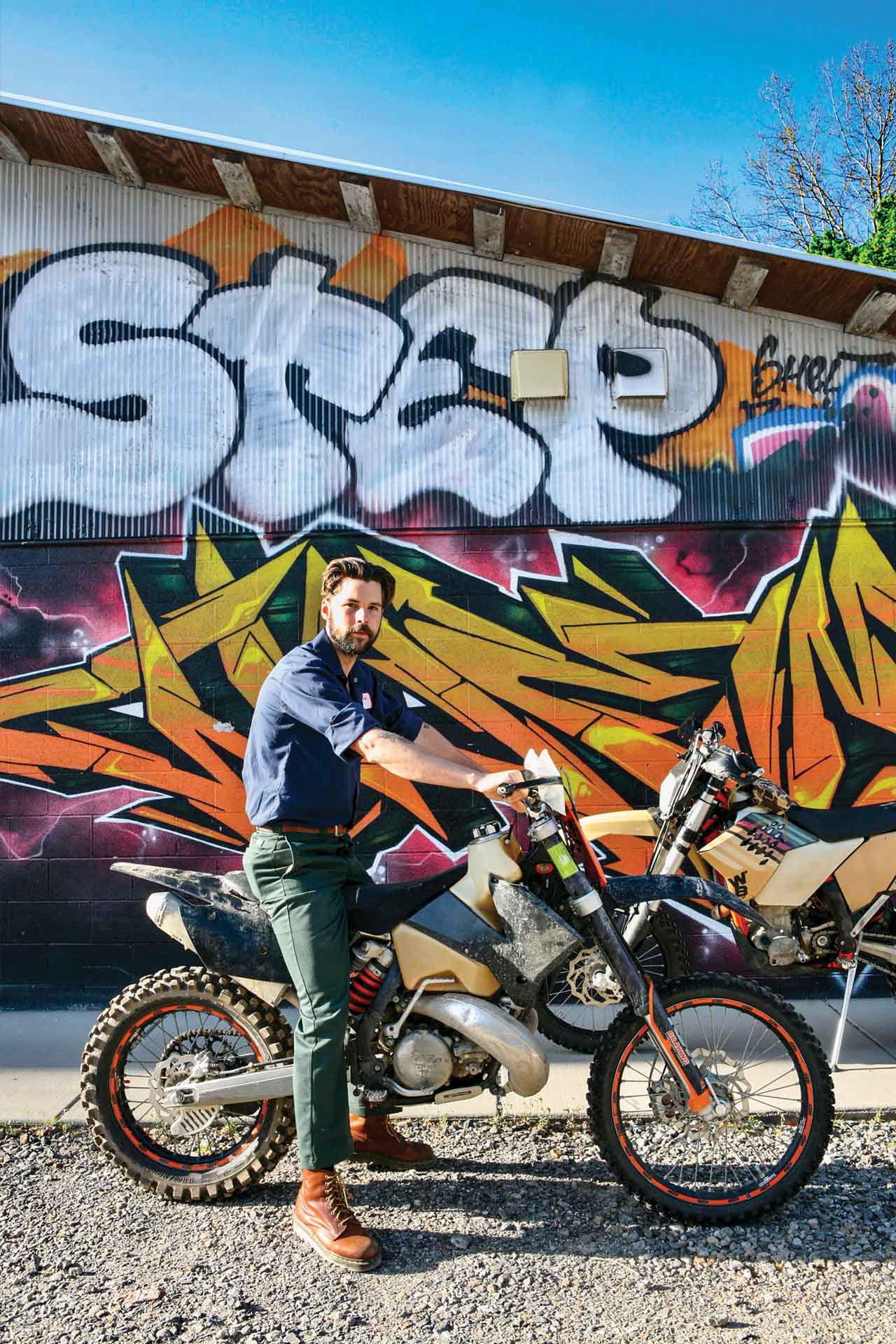 A man sits astride a vintage dirtbike in front of a colorful graffiti mural